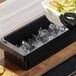 A black 4-compartment condiment bar with a clear lid, partially filled with ice and being stocked with lime wedges.