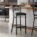 A black metal bar stool with a cross back design and a black vinyl padded seat.