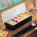 A six-compartment condiment bar filled with various garnishes such as lime slices, cherries, grapefruit, orange slices, and olives.