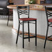 A black metal bar stool with a cross back design and a mahogany wood seat.