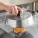 A person holding an American Metalcraft aluminum basting lid over a hamburger on a grill.