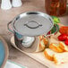 An American Metalcraft stainless steel pot with a lid on a cutting board next to a plate of bread and vegetables.