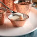 A close-up of a Choice hammered copper stainless steel sauce cup with a spoon pouring white liquid over a bowl of food.
