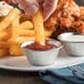 A hand dipping a french fry into a Choice stainless steel round sauce cup.