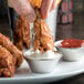 A person dipping a chicken wing into small stainless steel cups of sauce.