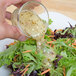 A hand pouring Ken's Foods Greek Vinaigrette with Feta Cheese and Black Olives dressing onto a bowl of salad.