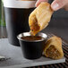 A hand holding a piece of food dipping it into a Choice matte black stainless steel round sauce cup.