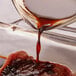 A close-up of Worcestershire sauce being poured from a glass container onto a piece of raw meat.