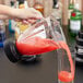 A person pouring a red liquid from a green bottle into a Waring commercial blender with a glass on the counter.