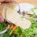 A hand pouring Ken's Foods Honey Mustard Dressing over a salad with carrots and onions.