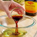 A hand pouring Maggi liquid seasoning from a small glass bowl into a larger glass mixing bowl.