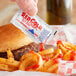 A hand holding a Red Gold ketchup packet over a burger.