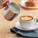 A person using a Choice stainless steel frothing pitcher to pour milk into a cup of coffee.
