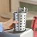A perforated stainless steel flatware holder cylinder containing utensils.