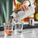 A person using an Acopa stainless steel cobbler cocktail shaker to pour a drink into a glass.