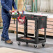 A man in blue overalls holding a black Rubbermaid utility cart.