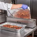 A person in a white coat and blue gloves using a stainless steel film and foil dispenser to cover a tray of food.