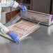 A person in a blue coat uses the Choice stainless steel film and foil cutter to cut plastic wrap over a tray of bacon.