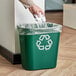 A hand throwing a crumpled paper into a green Rubbermaid recycling bin.