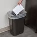 A hand putting a piece of paper into a Rubbermaid trash can.