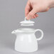 A hand holding a lid over a white Arcoroc teapot.