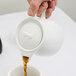 A person pouring brown liquid from an Arcoroc teapot into a cup.