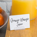An American Metalcraft white mini chalk card on a table with a glass of orange juice.
