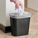 A hand placing crumpled white paper into a black Rubbermaid trash can.