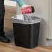 A hand putting a coffee cup into a Rubbermaid black rectangular wastebasket.