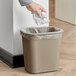 A hand holding a piece of paper and putting it into a Rubbermaid beige rectangular trash can with a plastic bag inside.