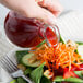 A hand pouring red liquid into a Tablecraft Olive Oil bottle on a table with a salad.