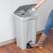 A woman in jeans and boots opens a Lavex Gray rectangular step-on trash can with her foot.
