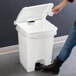 A woman opening a Lavex white rectangular step-on trash can with a plastic bag in it.