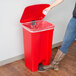 A woman using a Lavex red rectangular step-on trash can to put a plastic bag in.