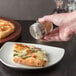 A hand using a Tablecraft spice shaker to sprinkle seasoning on a pizza.