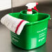 A green San Jamar Kleen-Pail container with a spray bottle and a towel.