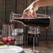 A hand pouring red wine from a Frilich glass carafe into a glass on a table.