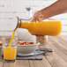 A hand pouring orange juice from a Frilich Tritan plastic carafe into a glass.