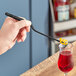 A hand holding a Mercer Culinary black offset tip plating tong over a glass of red liquid with a spiral.