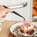 A hand using black Mercer Culinary curved tip tongs to plate a dessert with a pink butterfly on top.