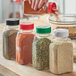 A person pouring spices into a rectangular plastic spice container with a black dual flapper lid with 3 holes.