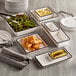 A table with a rectangular white and black melamine serving bowl full of food, with a stack of rectangular white plates and a spoon.