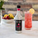 A close up of a Monin raspberry flavor concentrate bottle next to a glass of raspberry flavored beverage.