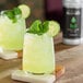 A glass of yellow liquid with ice and cucumber and a black bottle of Monin Basil Concentrated Flavor on a table.