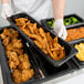 A person in white gloves holding a Cambro black plastic food pan full of food.