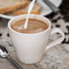 A close up of a Tuxton Europa white china cappuccino mug with a spoon being poured into a cup of coffee with milk.