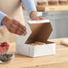 A white, customizable 6 x 6 x 2.5 inch bakery box being assembled on a wooden counter.