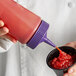 A person pouring sauce from a Vollrath Clear Squeeze Bottle with a purple cap into a cup of food.