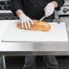A person in a white shirt and gloves cutting a loaf of bread with a San Jamar white cutting board and a knife.
