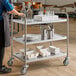 A stainless steel three-shelf utility cart holding glass bottles, glasses, plates, utensils, takeout containers, and napkins.